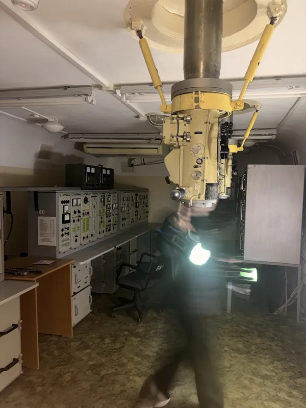 A control room with a periscope hanging from the ceiling and consoles along the wall.