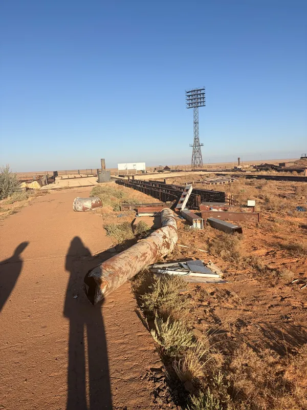 Scattered wreckage and a toppled pole on red earth, a floodlight mast behind.
