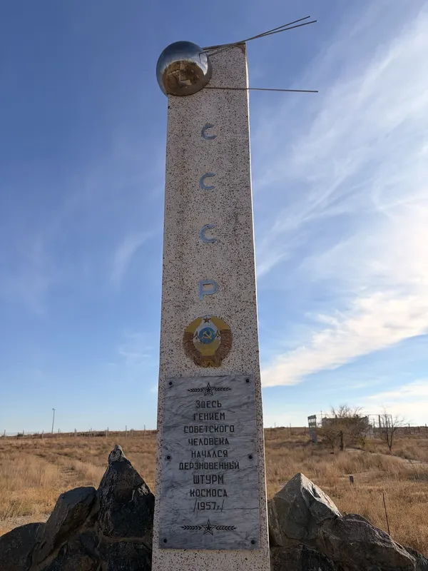A tall white obelisk topped with a model of Sputnik, with a marble plaque at its base.