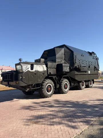 A dark eight-wheeled military transporter parked on a plaza.