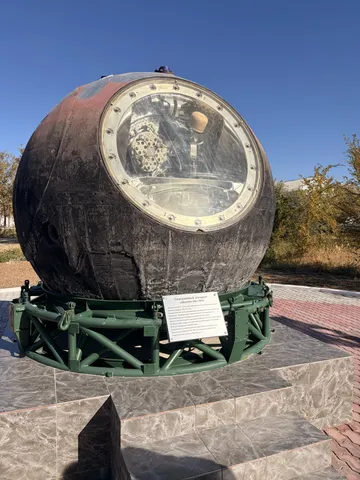 A charred spherical re-entry capsule with a round window, on a stand.